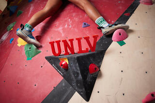 A cllimbers legs as they boulder their way up a climbing wall. Miniature UNLV letters are balanced on a climbing hold, between the climbers legs.